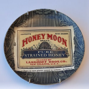 May include: A vintage Honey Moon Brand pure strained honey label, with text and a beehive illustration, is displayed on a decorative plate with a blue and gray floral design. The label reads "Packed by Langhoff Bros. Co., New Orleans, U.S.A."