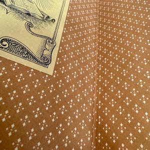 May include: Open book with a detailed illustration on a cream-coloured page. The book's interior features a brown patterned design with white diamond-shaped accents. The illustration depicts a ship, books, and a sun.