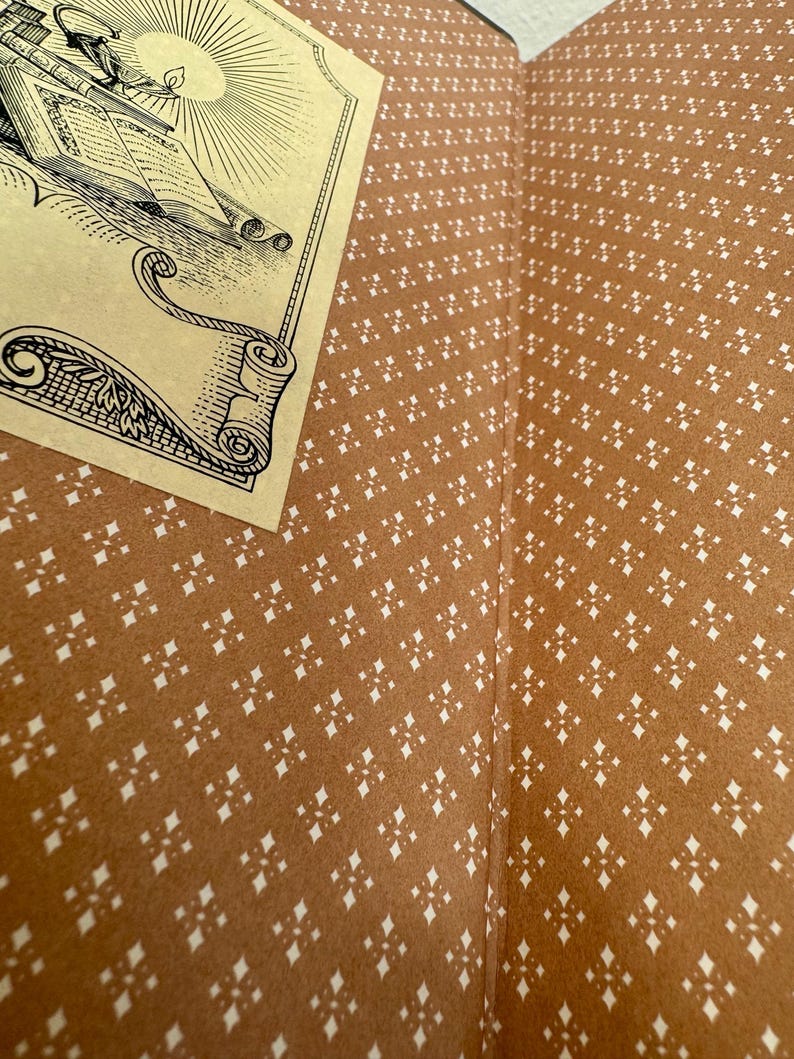 May include: Open book with a detailed illustration on a cream-coloured page. The book's interior features a brown patterned design with white diamond-shaped accents. The illustration depicts a ship, books, and a sun.