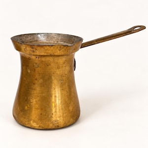 May include: A brass coffee pot with a wide, flared rim and a long, slender handle. The exterior surface of the pot shows a weathered, golden-brown patina with visible spots and marks, while the interior has a metallic, silver-toned finish.