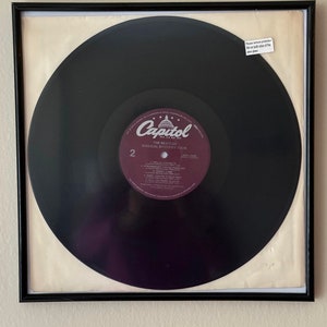 May include: A black vinyl record in a black frame with a white mat. The record is labeled "The Beatles Magical Mystery Tour" with the Capitol Records logo. The record is in a protective sleeve with a label that says "Please remove protection film on both sides of the  vinyl record."