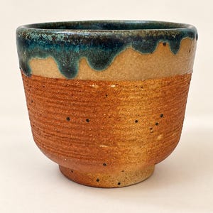 May include: A ceramic cup with a textured, reddish-brown base featuring horizontal ridges and small dark speckles. The upper portion of the cup is finished with a smooth, glossy teal glaze that drips down over the rim onto the textured surface.