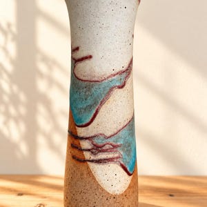 May include: A tall, cylindrical ceramic vase with a speckled off-white base and a flared rim. The surface features abstract, flowing patterns in shades of turquoise and deep maroon, with a textured, sandy-brown finish at the bottom section.