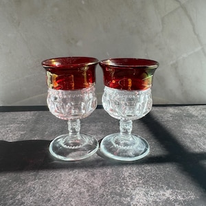 May include: Two vintage wine glasses with ruby red rims and clear, patterned bowls. The stems and bases are also clear. The glasses are displayed on a dark, textured surface, showcasing their elegant design.
