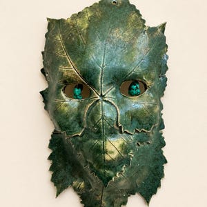 May include: A wall-mounted mask crafted from overlapping green and gold leaves. The mask features two eye openings, each accented with a small, bright turquoise stone. The surface displays detailed leaf veins and a textured, layered appearance.