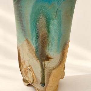 May include: A tall, cylindrical ceramic tumbler featuring a reactive glaze with a vibrant turquoise top that transitions into a textured, earthy beige base. The surface displays organic drips and variations in color, with a sturdy, slightly flared rim.