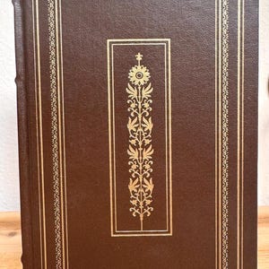 May include: A hardback book with a brown leather cover and gold detailing. The cover has a gold floral design within a rectangular frame, with a decorative border. The spine is also brown.