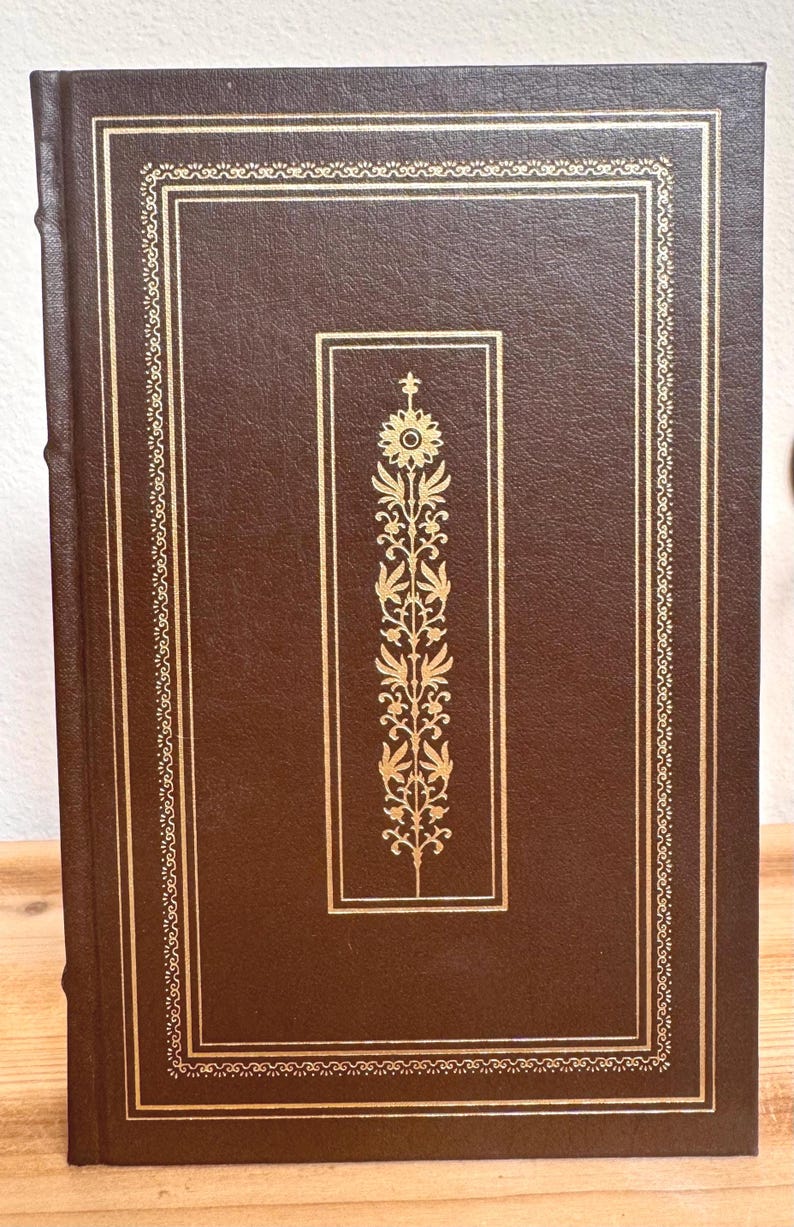 May include: A hardback book with a brown leather cover and gold detailing. The cover has a gold floral design within a rectangular frame, with a decorative border. The spine is also brown.