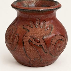 May include: A small, round, reddish-brown ceramic vase with a wide, flared rim. The exterior features a textured, dark brown embossed figure of a flute player, known as Kokopelli, flanked by two spiral patterns on the curved body of the vessel.
