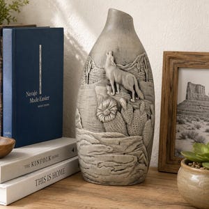 May include: A gray ceramic vase with a desert landscape design. The vase features a howling wolf, cacti, and mountains. The vase is displayed on a wooden surface with books and a framed photograph.