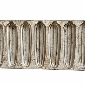 May include: A vintage, rectangular, silver-colored cast iron cornbread mold. The mold features six individual, elongated cavities, each with vertical ridges. The mold has a handle on each end and a hole for hanging.