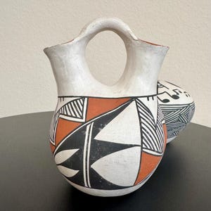 May include: A white pottery water jar with a handle and a rounded body. The jar is decorated with geometric patterns in black, orange, and white. A second, smaller jar with similar patterns is partially visible in the background.