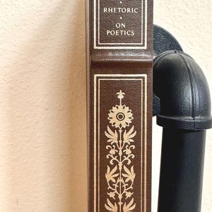 May include: A brown book titled "Aristotle Rhetoric on Poetics" with gold lettering and decorative floral patterns. The book is positioned vertically, with a black pipe visible on the right side. The book's spine is visible.