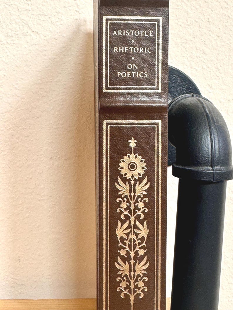 May include: A brown book titled "Aristotle Rhetoric on Poetics" with gold lettering and decorative floral patterns. The book is positioned vertically, with a black pipe visible on the right side. The book's spine is visible.