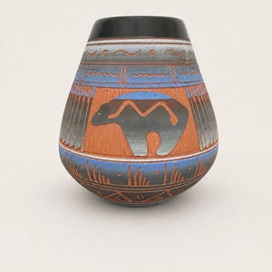 May include: A rounded ceramic vase featuring a black rim and a terracotta-colored body. The surface is decorated with a central dark gray bear silhouette, blue horizontal bands, and intricate etched geometric patterns and grass-like motifs.