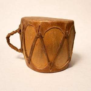May include: A small, handcrafted drum with a handle. The drum is made of light brown leather, with darker brown stitching and a tightly stretched drumhead. The handle is made of twisted leather.