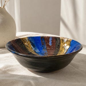 May include: A ceramic bowl featuring a dark black exterior and a vibrant interior glazed with vertical stripes of deep blue, metallic gold, and dark brown. The bowl has a wide, circular opening and a smooth, rounded base, showcasing a glossy finish.