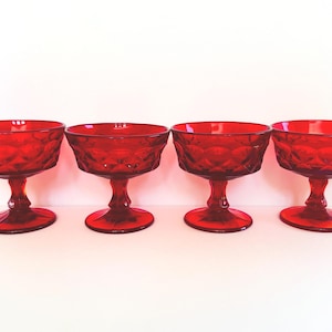 May include: Four ruby red glass dessert or champagne glasses. Each glass features a diamond pattern on the bowl and a pedestal base. The glasses are arranged in a row against a white background.