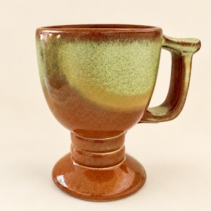 May include: A ceramic mug with a pedestal base and a wide handle. The upper portion features a mottled light green glaze, while the lower body, base, and handle are finished in a glossy, deep reddish-brown glaze with subtle tonal variations.