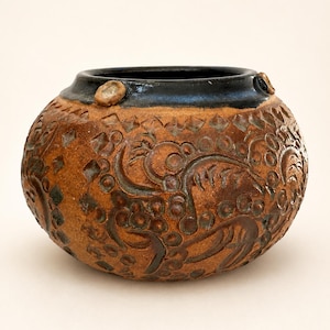 May include: A round, wide-mouthed ceramic pot with a textured, earthy brown surface featuring intricate, carved relief patterns of swirling shapes and geometric motifs. The rim is finished in a smooth, dark, glossy black glaze.