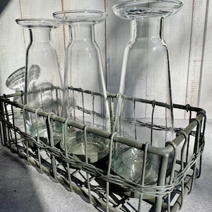 May include: Three clear glass vases with flared tops sit inside a gray metal wire basket. The vases have a slender, bottle-like shape. The basket has a rectangular form with a woven wire design. The background is a white wooden wall.