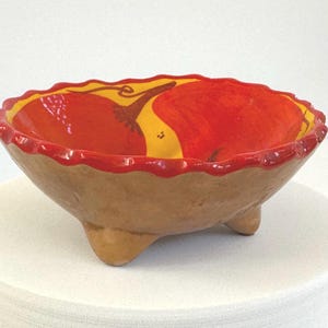 May include: A handmade, three-legged ceramic bowl. The interior features a vibrant red and yellow design. The rim is painted red, and the exterior is a natural brown color. The bowl is likely used for serving food or as a decorative piece.