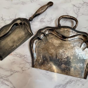 May include: Antique silver-toned crumb set, featuring a handled tray and a matching dustpan with a wooden handle. The tray has a curved handle and a decorative edge. The dustpan has a long handle and a decorative design.