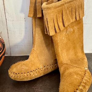 May include: A pair of tan suede boots with fringed tops. The boots have a moccasin-style toe and are stitched with a contrasting thread. The boots are knee-high and appear to be made of soft suede material.