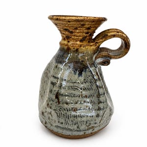 May include: A handmade ceramic pitcher with a handle. The pitcher has a bulbous body and a flared rim. The colors are primarily brown, gray, and blue, with a textured surface. The pitcher is likely used for serving beverages.