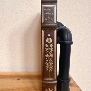 May include: A brown hardcover book titled "Aristotle Rhetoric on Poetics." The spine features gold lettering and decorative floral designs. The book is displayed vertically on a wooden surface.