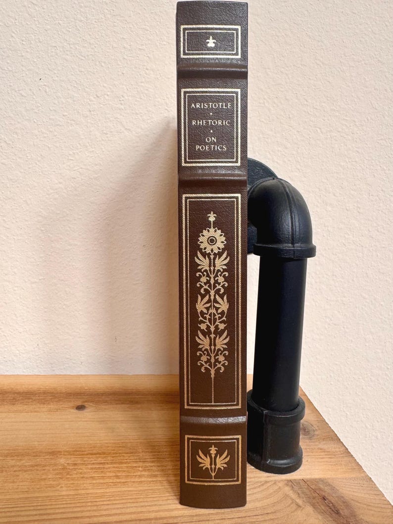 May include: A brown hardcover book titled "Aristotle Rhetoric on Poetics." The spine features gold lettering and decorative floral designs. The book is displayed vertically on a wooden surface.