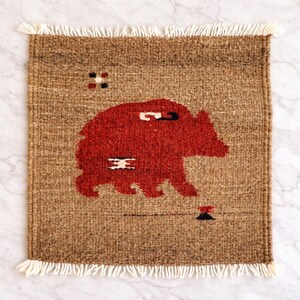 May include: A woven brown rug with a red bear design. The rug has a rectangular shape with a fringe border. The bear is the central design element, with a simple, stylized form. The rug is likely handmade.