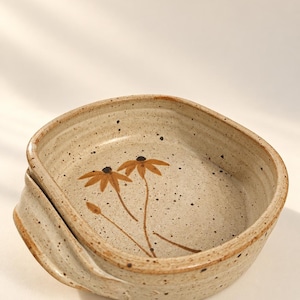 May include: A square ceramic bowl with rounded corners and integrated side handles. The bowl features a speckled cream glaze with a hand-painted design of two brown flowers with thin stems and a small bud on the interior base of the vessel.