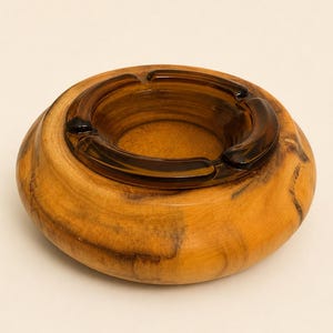 May include: A round ashtray featuring a polished natural wood exterior with visible dark grain patterns and a smooth, circular amber-colored glass insert. The glass center includes three integrated notches along the rim for resting items.