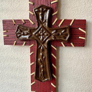 May include: A decorative cross with a red wooden base and a brown ceramic overlay. The red wood has a textured finish and is stitched together with cream-colored thread. The ceramic cross features floral and geometric designs.