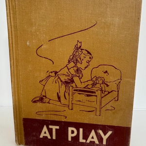 May include: Vintage book titled "AT PLAY" with a tan cover and maroon text. The cover features a maroon illustration of a girl playing with a doll in a bed. The book has a worn appearance, suggesting it is an antique.