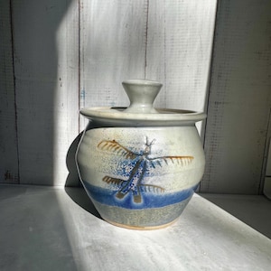 May include: A ceramic jar with a lid, featuring a dragonfly design. The jar is primarily beige with a blue band and dragonfly illustration. The lid has a small handle. The background is a white wooden surface.