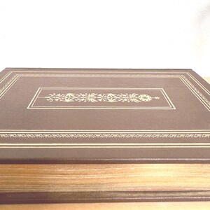 May include: A brown, rectangular book with a decorative gold floral design in the centre and along the edges. The book's cover has a textured appearance, and the pages are visible along the side.