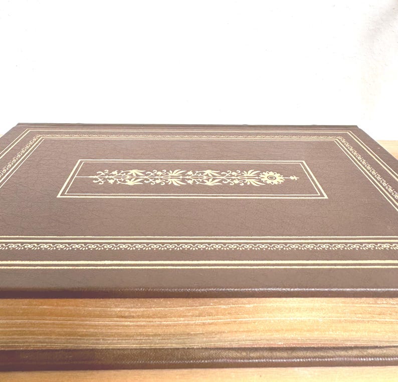 May include: A brown, rectangular book with a decorative gold floral design in the centre and along the edges. The book's cover has a textured appearance, and the pages are visible along the side.