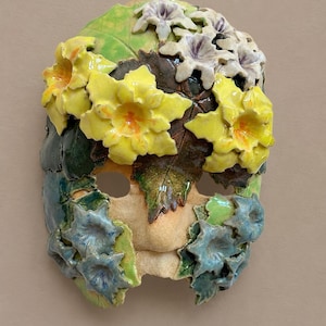 May include: A ceramic mask decorated with three-dimensional sculpted flowers and leaves. The top features white and yellow blossoms, while the lower section displays blue flowers. The base is a textured, light-colored clay with green foliage.