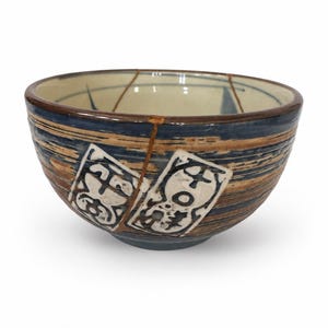 May include: A ceramic bowl featuring a dark blue and brown horizontal striped pattern with two white rectangular panels containing black abstract symbols. A thin, gold-colored line runs vertically down the side, indicating a repair.