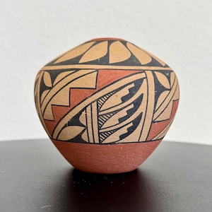 May include: A small, round, hand-painted pottery vessel with a reddish-brown base and a beige, red, and black geometric design. The top features a pattern of black and beige leaf shapes.