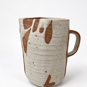 May include: A cylindrical ceramic mug with a textured, ribbed surface. The mug features a raw, reddish-brown clay body partially covered by a thick, off-white glaze that drips down the sides, revealing the natural clay texture underneath.