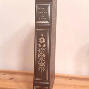 May include: A dark brown hardcover book with gold lettering and decorative gold floral patterns on the spine. The title "ARISTOTLE RHETORIC ON POETICS" is displayed. The book has a small emblem at the top and bottom.