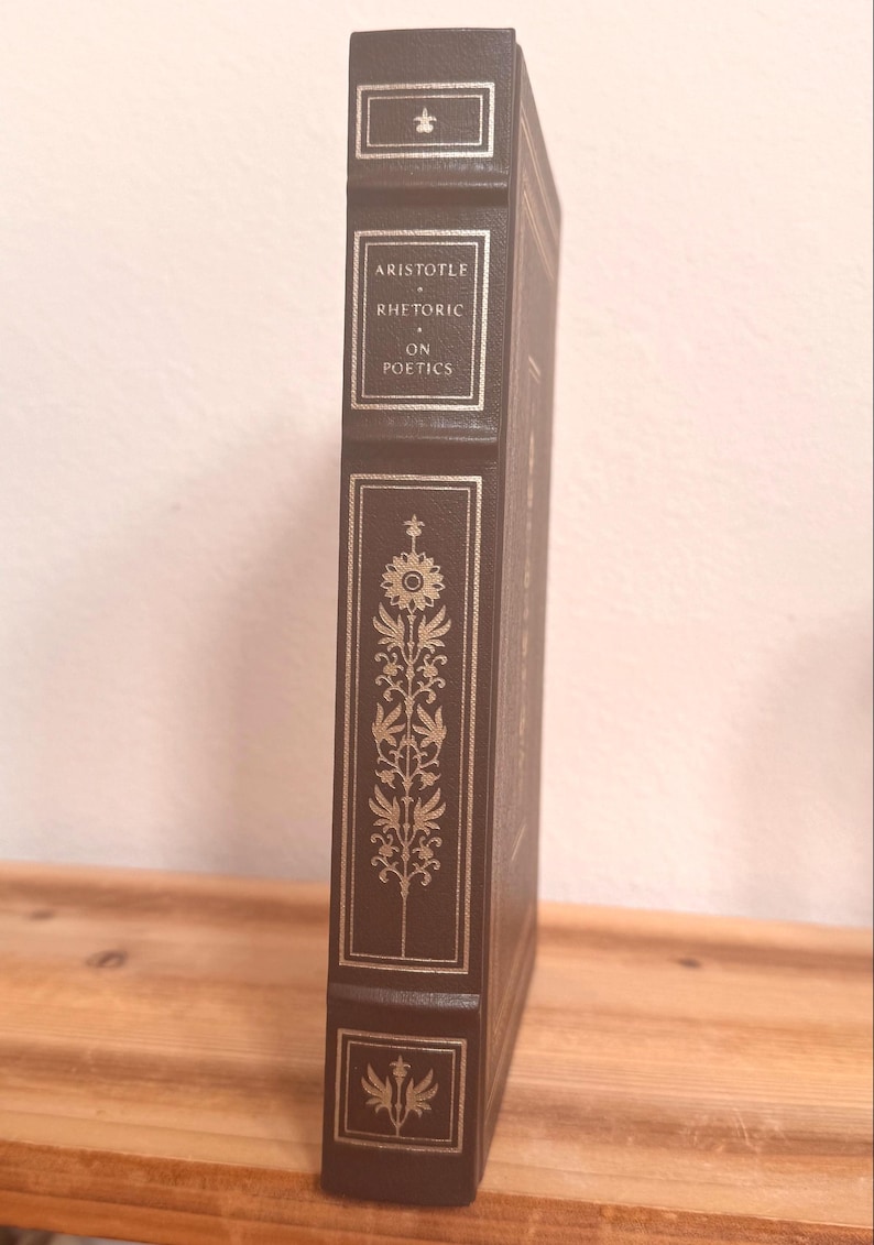 May include: A dark brown hardcover book with gold lettering and decorative gold floral patterns on the spine. The title "ARISTOTLE RHETORIC ON POETICS" is displayed. The book has a small emblem at the top and bottom.