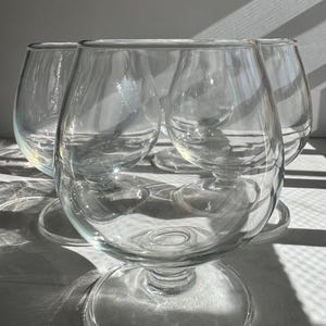 May include: Four clear, stemmed brandy snifters are arranged in a group. The glasses have a rounded bowl shape and sit on a circular base. The light reflects off the glass, creating shadows.