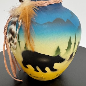 May include: A hand-painted ceramic vase with a bear silhouette, mountains, and trees. The vase features a gradient of yellow, blue, and green colors. It is adorned with feathers and a braided leather strap around the rim.