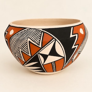May include: A rounded ceramic bowl featuring a cream-colored base with intricate hand-painted geometric patterns in matte black and terracotta orange. The design includes sharp zig-zag lines, circular motifs, and star-like shapes.
