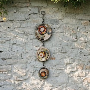 May include: A wooden garden bench with two patterned cushions, a small metal watering can, and three circular decorative wall hangings featuring colorful mosaic patterns, suspended vertically by a metal chain against a stone wall.