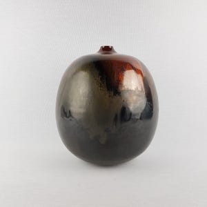 May include: A spherical ceramic vase with a narrow opening at the top. The surface features a glossy, multi-toned glaze transitioning from deep black at the base to metallic bronze and dark red hues near the top, creating a reflective finish.